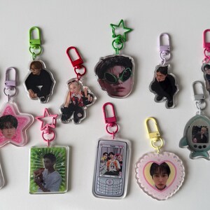 May include: A collection of thirteen acrylic keychains featuring various shapes including a slice of bread, stars, hearts, a camera, a mobile phone, and a fish, each with a colourful clasp and printed with photographic images of people.