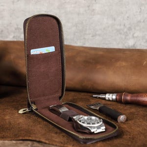 Pode incluir: Um estojo de relógio de couro marrom com zíper, aberto para revelar um relógio de pulso prateado com uma pulseira de couro marrom. O estojo também tem um pequeno bolso para um cartão. O fundo inclui couro e ferramentas.