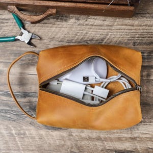 May include: A tan leather pouch with a zip, open to show white electronic accessories. The pouch has a handle and is on a wooden surface. Green and silver pliers are visible in the background.