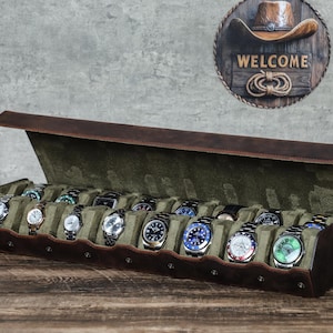 May include: A brown leather watch case with a green interior, holding a collection of various wristwatches. The case is open, displaying the watches. A wooden sign with a cowboy hat and the word "WELCOME" hangs in the background.
