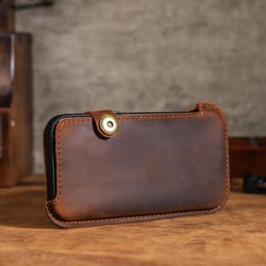 May include: A brown leather phone case with a snap closure. The case is rectangular with a stitched border and a brass-colored snap. The leather has a distressed appearance, and the case appears to be designed to hold a smartphone.