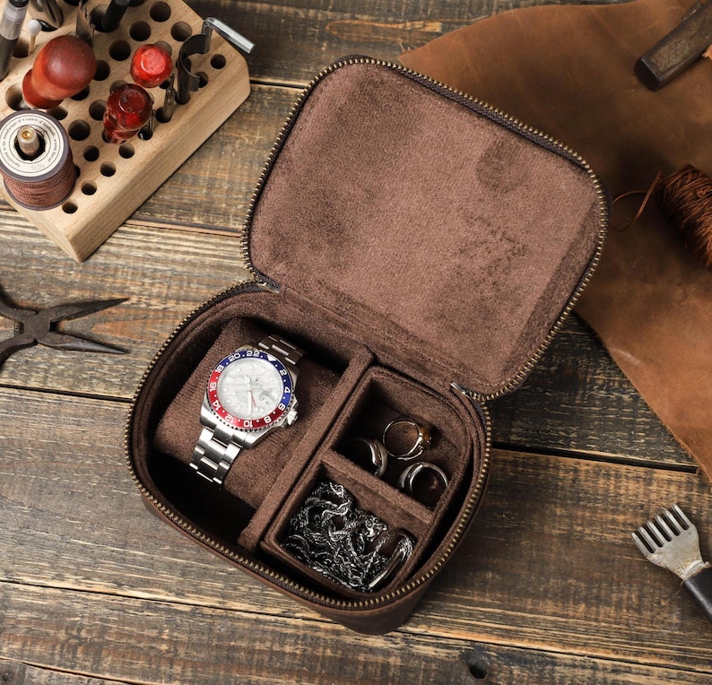 May include: A brown leather watch travel case with a zipper closure. The case is open and a silver watch with a white face and red and blue accents is inside. There are also two small compartments for rings or other jewelry.