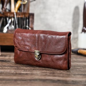 May include: A brown leather clutch with a flap closure and a metal clasp. The clutch is rectangular and appears to be made of high-quality leather. The background includes tools, suggesting a handcrafted item.