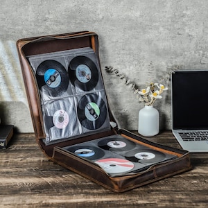 Puede incluir: Estuche de CD de cuero marrón abierto con cremallera, lleno de discos de vinilo. El estuche está sobre una superficie de madera junto a un portátil y un pequeño jarrón con flores. Los discos tienen etiquetas de varios colores.