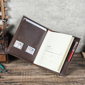 Puede incluir: Portafolio de cuero marrón abierto con bolsillo para tableta, dos ranuras para tarjetas, un bloc de notas y un bolígrafo rojo. El bloc de notas tiene las palabras "PERSONAL DATA" en inglés y chino.