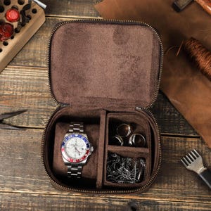 May include: A brown leather travel case with a zipper closure. The case is open and contains a silver watch with a blue, red, and white face, a small compartment with rings, and a larger compartment with a silver chain.
