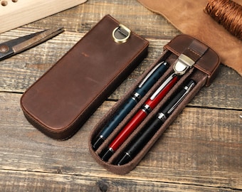 May include: A brown leather pen case with a metal clasp. The case is open and contains three pens, one blue, one red, and one black.