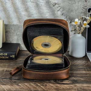 May include: A brown leather CD case, open to show gold CDs in clear sleeves. The case has a zip closure and a small handle. The background includes a book, a vase with flowers, and a laptop.