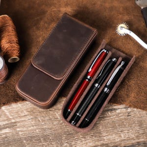 May include: A brown leather pen case with three slots for pens. The case is open and three pens are visible: one red pen and two black pens.