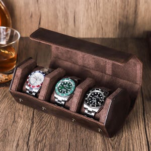 May include: A brown leather watch case containing three wristwatches with silver straps and coloured faces. The faces are red, green, and black. A glass of amber liquid and a bottle are in the background.