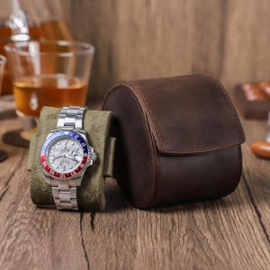 Puede incluir: Un rollo de reloj de cuero marrón con un reloj plateado con una esfera blanca y un bisel rojo y azul. El reloj está sobre un cojín de terciopelo verde.