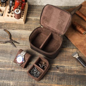 May include: A brown leather watch box with a zipper closure. The box has two compartments for storing watches and a separate compartment for storing jewelry. The box is open and the compartments are lined with brown velvet.