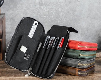 May include: A black zippered leather pen case with an open interior containing four pens, a small white rectangular eraser, and a metal ruler. A stack of four additional closed pen cases in red, green, gray, and brown sits to the right.