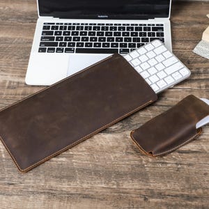 Könnte beinhalten: Ein braunes Leder-Tastatur-Etui und eine Maus-Hülle liegen auf einem Holztisch neben einem Laptop. Das rechteckige Tastatur-Etui und die kleinere Maus-Hülle sind aus Leder mit sichtbaren Nähten gefertigt.