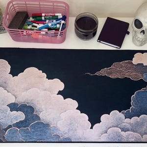 May include: A rectangular desk mat featuring a dark navy blue background with stylized, layered clouds in shades of light pink, white, and muted blue. The cloud patterns include intricate, fine-lined circular textures throughout the design.