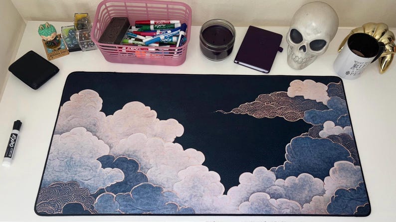May include: A rectangular desk mat featuring a dark navy blue background with stylized, layered clouds in shades of light pink, white, and muted blue. The cloud patterns include intricate, fine-lined circular textures throughout the design.