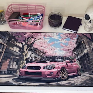 May include: A rectangular desk mat featuring a printed illustration of a pink sports car parked on a street lined with traditional buildings and cherry blossom trees. The mat has a black stitched border and a smooth surface for computer mouse use.