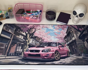 May include: A rectangular desk mat featuring a printed illustration of a pink sports car parked on a street lined with traditional buildings and cherry blossom trees. The mat has a black stitched border and a smooth surface for computer mouse use.