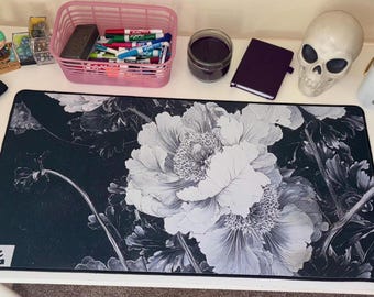 May include: A rectangular desk mat featuring a high-contrast black and white floral pattern with large, detailed peony-like blossoms. The mat has a black background, white petals, and a small white square in the bottom left corner with black text.