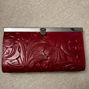 Op de afbeelding: Een rode leren portemonnee met een reliëf bloemenontwerp. De portemonnee heeft een zilverkleurige sluiting en een decoratief metalen accent. De portemonnee is rechthoekig en lijkt gesloten.