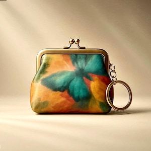 May include: A colorful coin purse with a kiss-lock closure and a key ring. The purse features a vibrant watercolor design in shades of green, orange, and yellow. The metal frame and key ring have a gold finish.