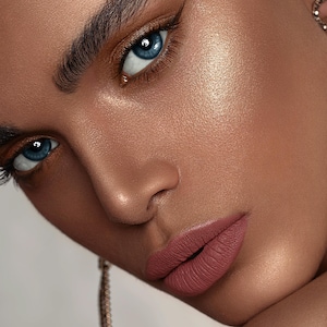 May include: Close-up of a person's face with blue eyes, defined eyebrows, and a soft pink lipstick. The skin has a natural glow and is highlighted with a shimmery product.