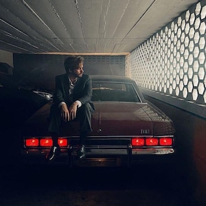 May include: A vintage red Ford sedan with illuminated taillights. A person in a dark suit sits on the car's trunk. The scene is set in a dimly lit garage with a honeycomb-patterned wall.