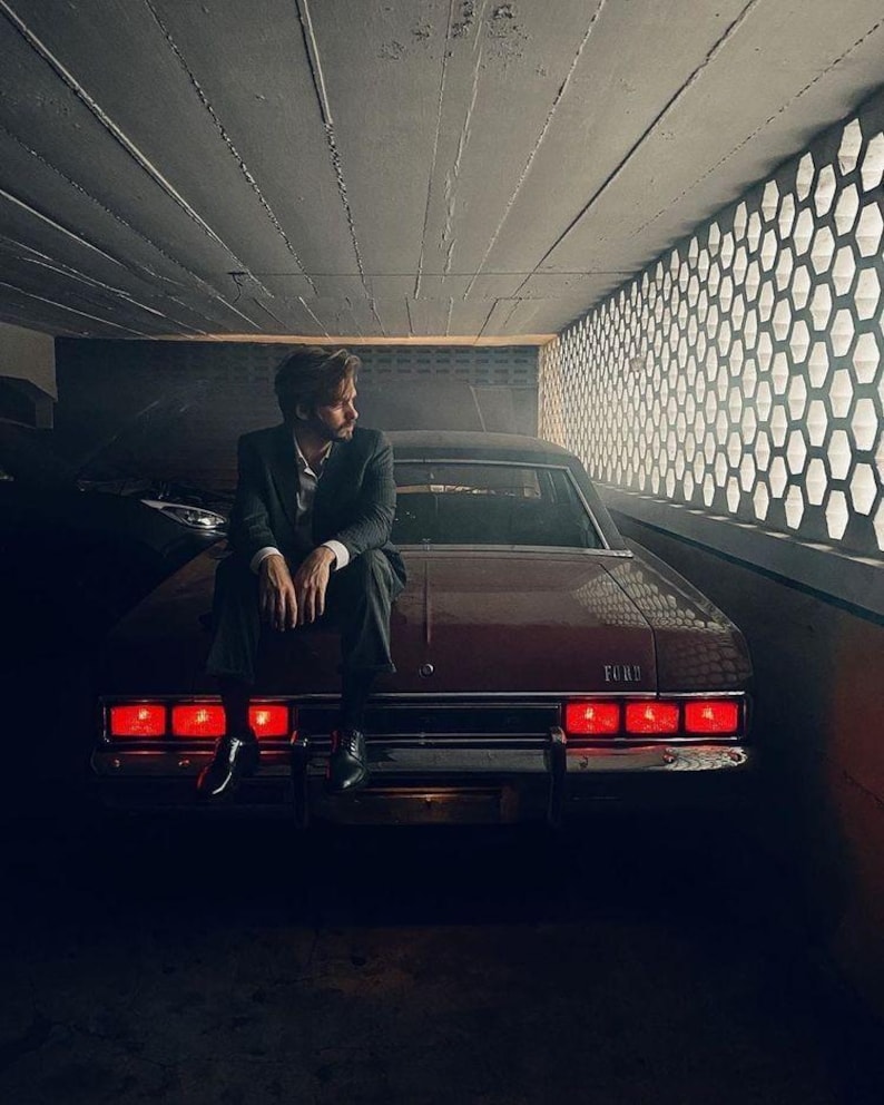 May include: A vintage red Ford sedan with illuminated taillights. A person in a dark suit sits on the car's trunk. The scene is set in a dimly lit garage with a honeycomb-patterned wall.