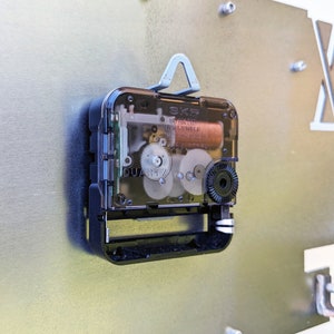 May include: Close-up of a clock mechanism with a clear plastic cover. The mechanism is silver and black with visible gears and a small battery. The text "SKP" and "QUARTZ" are visible on the mechanism.