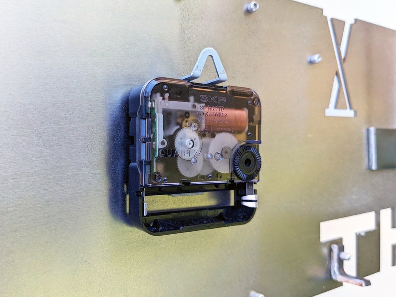 May include: Close-up of a clock mechanism with a clear plastic cover. The mechanism is silver and black with visible gears and a small battery. The text "SKP" and "QUARTZ" are visible on the mechanism.