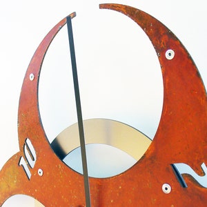 May include: A rusty metal clock with Roman numeral markings. The clock face is made of three overlapping crescent shapes. The clock hands are black.