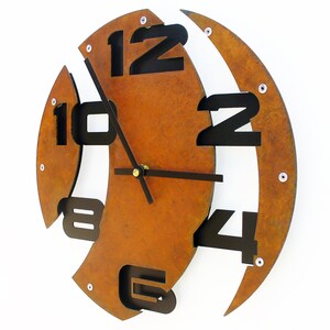 May include: A rusty brown metal wall clock with black numbers and hands. The numbers are arranged in a unique, asymmetrical pattern.