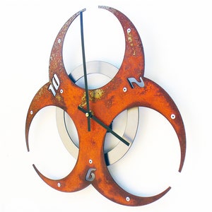 May include: A rusty orange metal wall clock shaped like a biohazard symbol with black clock hands and silver numbers.