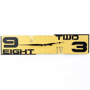 May include: A gold-colored clock with black Roman numeral markings and black numbers. The clock face is divided into two sections, with the left section displaying the number "9" and the word "EIGHT" and the right section displaying the number "3" and the word "TWO".