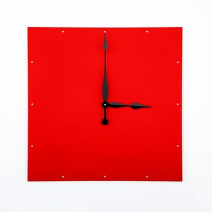 May include: A square wall clock with a vibrant red face and black hands. The clock's hands point to approximately 3 o'clock. The clock has small silver dots marking the hours. The clock is mounted on a white wall.