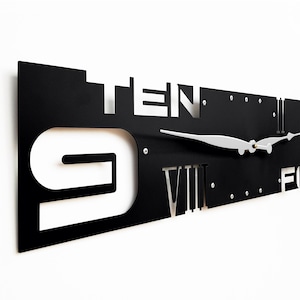 May include: Black metal wall clock with white Roman numeral and Arabic numeral hour markers. The clock hands are silver.