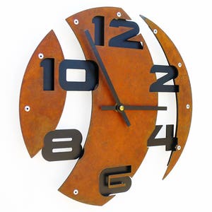 May include: A rusty orange metal wall clock with black numbers and hands. The numbers are arranged in a unique, asymmetrical pattern.