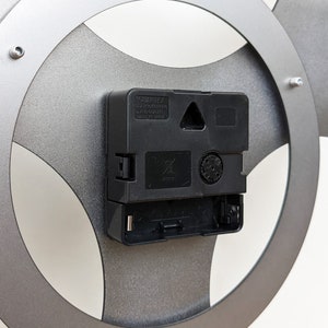 May include: The back of a silver clock with a black clock mechanism. The clock mechanism has a small black triangle and a square on it. The text "QUARTEX" is visible on the clock mechanism.