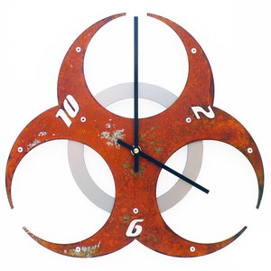 May include: A rusty red metal clock shaped like a biohazard symbol with black hands and white numbers. The clock has a silver inner circle.