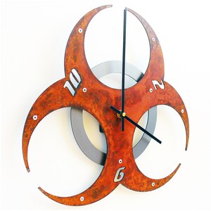 May include: A rusty orange metal wall clock shaped like a biohazard symbol with black hands and silver numbers.