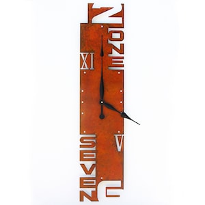 May include: A rectangular, rusty-orange metal wall clock with cut-out numerals and the words "ONE", "SEVEN", and "TWO". The clock has black hands and silver Roman numeral "XI". The clock is mounted on a white wall.