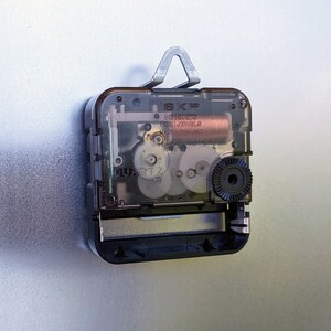 May include: Close-up of a black clock mechanism with a clear plastic cover. The mechanism has a silver metal hook on the top and a black knob on the right side. The text "SKIP 80 HR LTD MVT 12 JEWELS MADE IN THAILAND" is printed on the mechanism.