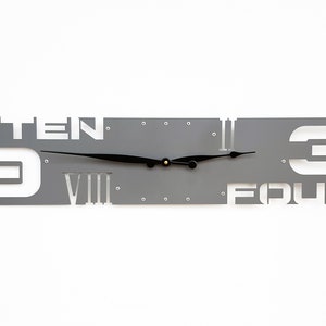May include: Gray metal wall clock with Roman numeral and number hour markers. The clock face is cut out to show the numbers "9", "TEN", "FOUR", and "3".