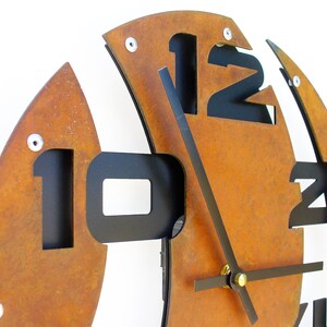 May include: A rusty brown metal clock with black numbers and hands. The clock is designed in a unique, abstract style with the numbers cut out of the metal.