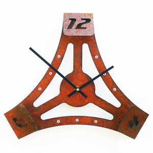 May include: A rusty metal clock with a triangular shape and black hands. The clock face is a circle with a gold center and the numbers 12, 3, and 6 are visible.