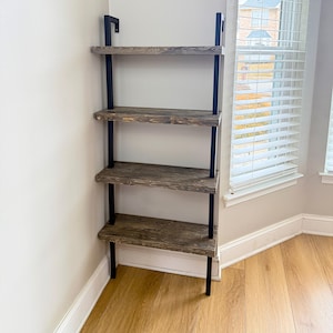 May include: A four-tier wooden shelf unit with black metal supports. The shelves are made of rustic wood with a dark brown finish. The shelf unit is mounted to a wall and is in a corner.