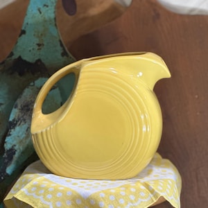 May include: A yellow ceramic pitcher with a rounded shape and a wide spout. The pitcher has a smooth, glossy finish and a subtle ribbing pattern.