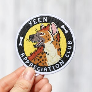 Op de afbeelding: Een ronde, zwart-gele geborduurde patch met een witte rand. De patch toont een cartoonhyena met bruin, oranje en wit bont. De tekst "YEEN APPRECIATION CLUB" is in wit rond de hyena geschreven.