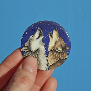 Könnte beinhalten: Ein runder Knopf mit blauem Hintergrund und weißen Sternen. Zwei Wölfe heulen zum Mond, einer weiß und einer braun. Die Wölfe stehen sich gegenüber.