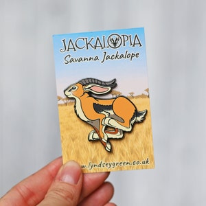 May include: A Jackalopia enamel pin featuring a cartoon jackrabbit with orange and black fur, running on a yellow background. The text "Jackalopia" and "Savanna Jackalope" are printed on the card. The website "w.lyndseygreen.co.uk" is printed on the bottom of the card.